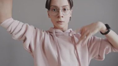 Gözlüklü ve açık pembe kapüşonlu bir gencin orta boy portresi. Gri duvarın önünde dans ediyor, kameraya bakıyor.