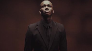 Yakışıklı, Afro-Amerikalı, siyah elbiseli, kahverengi arka planda dikilen ve yukarı bakan genç bir adamın stüdyo portresini kaldır.