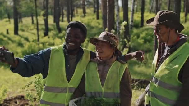 在阳光明媚的日子里 三名多种族生态志愿者在森林里用智能手机自拍树苗 — 图库视频影像