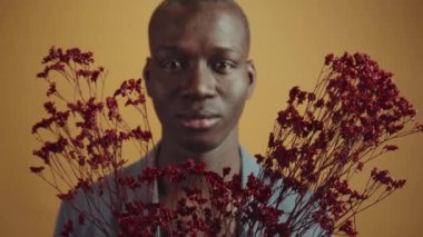 Seçici odaklı çekimde, elinde bir demet tutmakta olan Afrikalı Amerikalı bir genci veya soluk turuncu-sarı arka planda duran koyu kırmızı çiçekleri yakınlaştır.