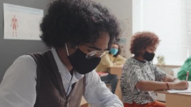 Maskeli ve gözlüklü zeki Afro-Amerikan adamın ders masasında oturması, öğleden sonra özet yazması.