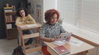 Afro-Amerikan kız öğrencinin tablet bilgisayar kullanarak okul masasında oturduğu orta boy portresi. Sonra yukarı bakıp kameraya gülümsüyor.