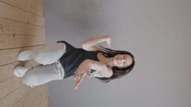 Gündüz vakti gri duvarın önünde dans eden siyah kolsuz tişörtlü ve eşofmanlı genç kadının dikey hâli. Kameraya bakıyor.