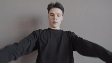 Siyah kazaklı genç bir adamın orta boy portresi. Gri duvarın önünde dans ediyor, kameraya bakıyor.