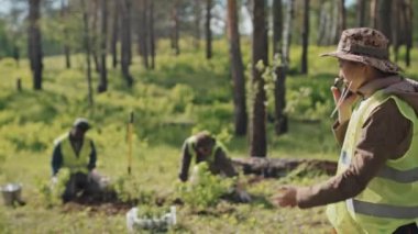 Orta boylu Asyalı kadın ormancı cep telefonuyla konuşuyor, güneşli bir yaz gününde ormanda duruyor.