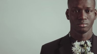 Siyah ceket ve çiçek desenli gömlek giyen, elinde kameraya bakan beyaz çiçeklerle şık Afrikalı Amerikalı adamın stüdyo portresi.
