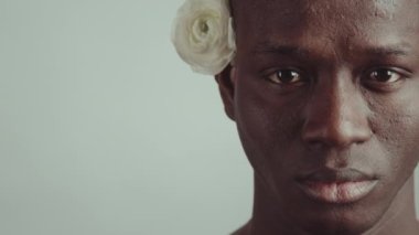 Kulağının arkasında taze beyaz gül çiçeği olan ve kameraya bakan genç Afro-Amerikan bir adamın yakın çekim portresi.