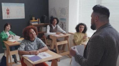 Erkek biyoloji öğretmeninin çeşitli öğrencilere ders vermesi, kıvırcık saçlı kızın el kaldırması, sabahları masa başında oturması.