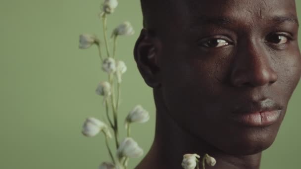 年轻黑人男子的特写 手持新鲜的白花 看着相机 苍白灰蒙蒙的绿色背景 — 图库视频影像