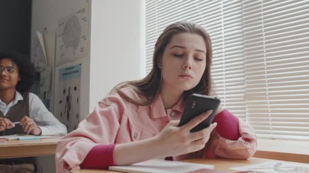 女性の低角度白人高校生は午前中に教室の机に座って スマートフォンを使用して — ストック動画