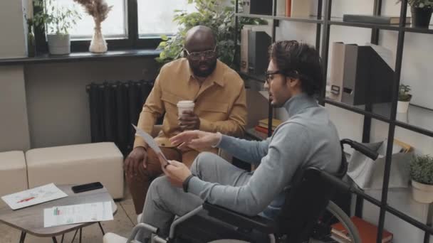 男性と話す車椅子の中近東の男コーヒー紙コップ付きの黒人同僚昼間はオフィスで書類を扱う — ストック動画
