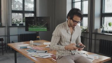 Gözlüklü genç Orta Doğulu bir adamın portresi öğleden sonra konferans masasında oturuyor, akıllı telefon kullanıyor, kameraya bakıyor ve gülümsüyor.