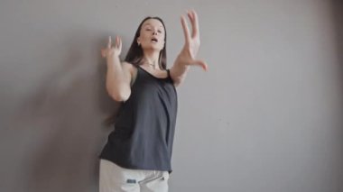 Gündüz vakti gri duvarın önünde dans eden siyah kolsuz tişörtlü, orta boylu, kahverengi saçlı bir kadın.