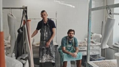 Kafkasyalı engelli bir adamın yavaş çekimde portresi ve erkek iş arkadaşı protez üretim atölyesinde dikilip kameraya bakıyor.