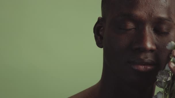 年轻的黑人男子手持白花 紧闭双眼 面对苍白的绿色背景 工作室特写 — 图库视频影像