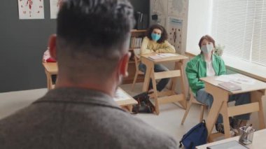 Erkek profesörün, yüz maskesi takan ve sabahları masalarında oturan genç öğrencilere anatomi dersi vermesine arka bakış.