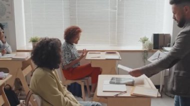 Masaların arasında yürüyen beyaz erkek öğretmenin gündüz vakti sınıftaki lise öğrencilerine kurbağa anatomisi haritası olan çarşafları verişi.