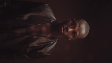 Deri ceketli özgüvenli Afrikalı Amerikalı adamın dikey orta boy portresi. Koyu kahverengi arka planda, disko topu aydınlatmalı.