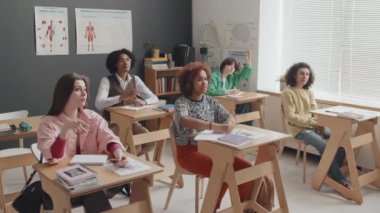 Orta uzunluktaki çok ırklı üniversite öğrencileri sınıfta masalarında oturup gündüz el kaldırıyorlar.