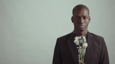 Siyah ceketli, çiçek desenli gömlekli, elinde çiçek buketi olan ve kameraya bakan genç Afro-Amerikan bir adamın stüdyo portresi.