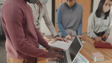 Portatif bilgisayar kullanan, gündüz vakti toplantı masasında maske takan, belgelerle uğraşan, konuşan çok ırklı iş arkadaşları.