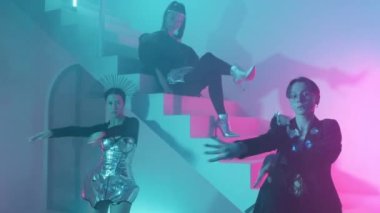 Siyah takım elbiseli ve korseli bir adam beyaz merdivenlerde oturuyor. Metalik detayları olan iki kız neon ışıklarla dans ediyor.