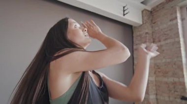 Siyah kolsuz tişörtlü genç bir kadının orta boy portresi. Gri duvarın önünde dans ediyor, kameraya bakıyor.