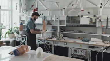 Önlük ve eldiven giyen genç beyaz bir adam modern atölyede bej sıvı plastikle protez bacak soketi detaylarını oluşturuyor.
