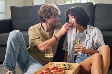 Genç ve sevecen bir adam, karısının ağzına küçük bir parça peynir koyarken her ikisi de kanepede otururken şampanya flütüyle...