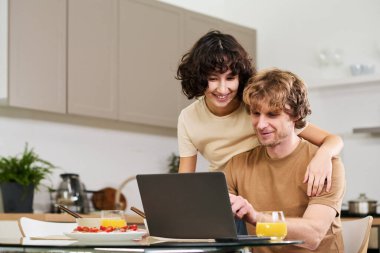 Hem laptop ekranına bakıp hem de kahvaltıda internetten mal seçen mutlu genç bir kadın.