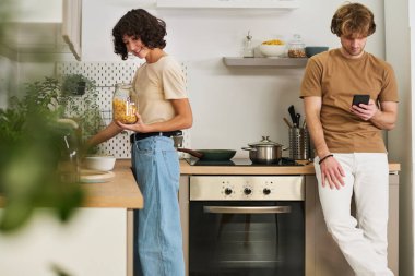 Lavabonun yanında dururken elinde mısır gevreği kavanozu tutan genç bir kadın ve akıllı telefonlu kocası kontakları veya internet şeylerini karıştırıyor.