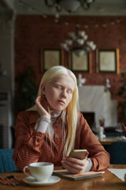 Uzun sarı saçlı, rahat albino kız kafede takılırken akıllı telefon ekranına bakıyor ve merak uyandıran online video dosyaları arıyor.