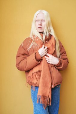 Uzun saçlı, mavi kotlu, kahverengi ceketli ve atkılı albino kız sarı duvara ya da arka planda tek başına dikilip yan yana bakıyor.