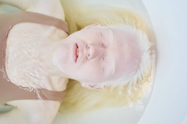 Temiz su dolu küvette uzanan ve fotoğraf çekimi sırasında dinlenen bej rengi tişörtlü genç bir albino kadın.