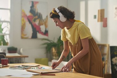 Tulumlu ve tişörtlü genç bir kadın ev stüdyosundaki yeni sanat şaheserinde çalışırken kulaklıkla müzik dinliyor.
