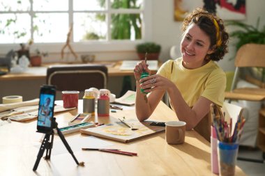 Resim fırçası ve guaj ile gülümseyen genç, yaratıcı bir kadın. İş yerinde oturuyor ve yüksek lisans sırasında akıllı telefon kamerasına bakıyor.