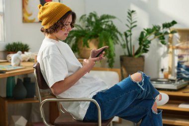 Rahat bir stüdyoda kameranın önünde otururken şık giyinen, akıllı telefonla mesajlaşan genç bir kadının yan görüntüsü.