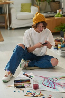Akıllı telefonlu genç yaratıcı kadın ressam yerde otururken onun tarafından kağıda boyanmış yeni sanat eserlerinin fotoğrafını çekiyor.