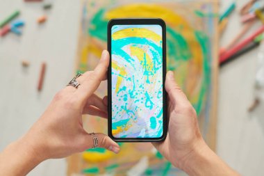 Genç yaratıcı kadın ressamın tuttuğu, mavi ve sarı suluboya ile boyanmış yeni soyut sanat eserlerinin fotoğrafını çeken akıllı telefon açısının üstünde.
