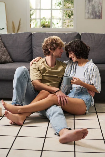 年轻快乐的一对光着脚的情侣 穿着便服 躺在沙发旁的客厅地板上放松 而黑发女孩则在喝咖啡 — 图库照片