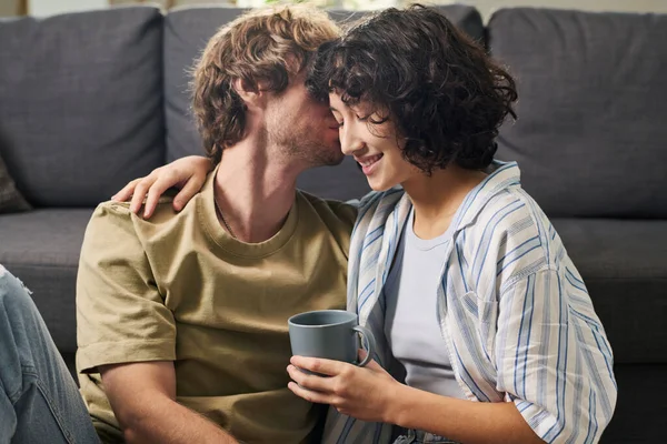 年轻的跨文化浪漫夫妇在客厅的沙发上享受着家的休息 而漂亮的女人则喝一杯咖啡拥抱着她的丈夫 — 图库照片