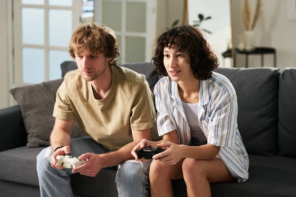 若いカップルでコントロールパッドを見ながらテレビセットの画面でリビングルームでソファに座ってレジャーやビデオゲームをプレイ — ストック写真