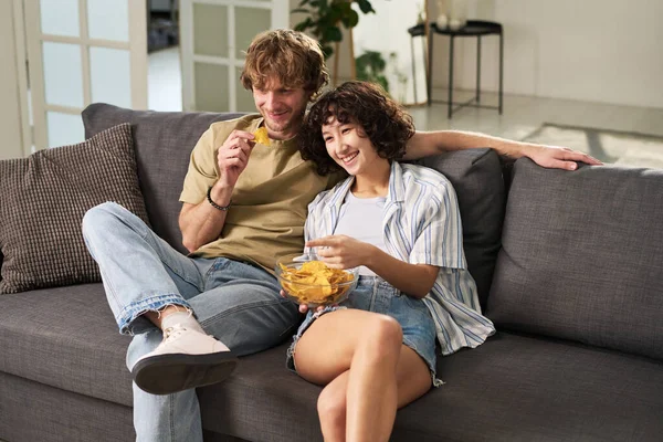 幸せな若いです夫と妻笑っている間にソファの上に座ってテレビセット 食べるポテトチップスと見てコメディにレジャー — ストック写真