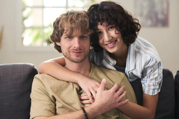年轻多情的夫妻在沙发上放松下来 看着相机 而快乐的黑发女人俯伏在丈夫身边拥抱他 — 图库照片