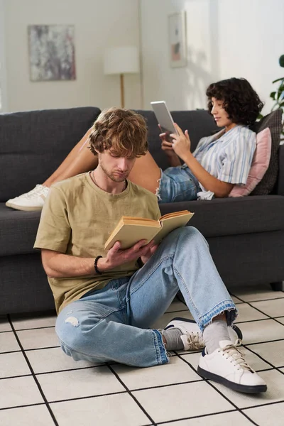 年轻而安详的男人躺在地板上看书 而他的妻子则带着平板电脑躺在沙发上休息 看在线电影或上网 — 图库照片