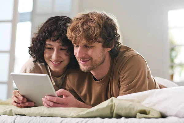 快乐的年轻夫妇们 早上睡懒觉后躺在床上休息 一边看有趣的网上视频或电影 一边吃平板电脑 — 图库照片