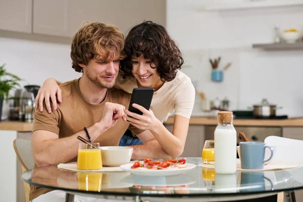 年轻的微笑的女人在智能手机里给她的丈夫展示一些东西 她坐在桌子旁边 一边吃三明治 果汁和牛奶 一边吃早饭 — 图库照片