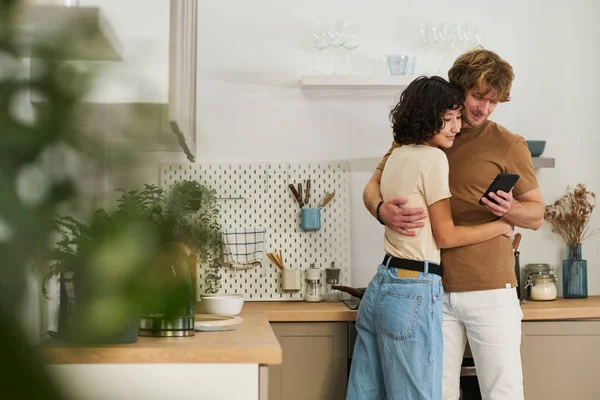 年轻女人拥抱着丈夫 站在厨房里看着男人拿着的智能手机屏幕 — 图库照片