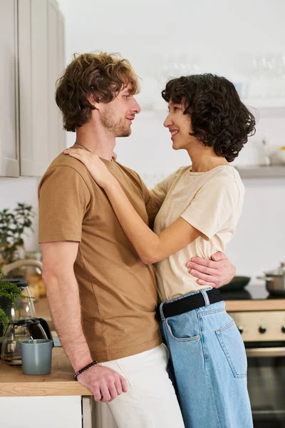 快乐的年轻夫妇穿着便服互相望着对方 而男人则拥抱着女人 把她的手放在他的肩上 — 图库照片
