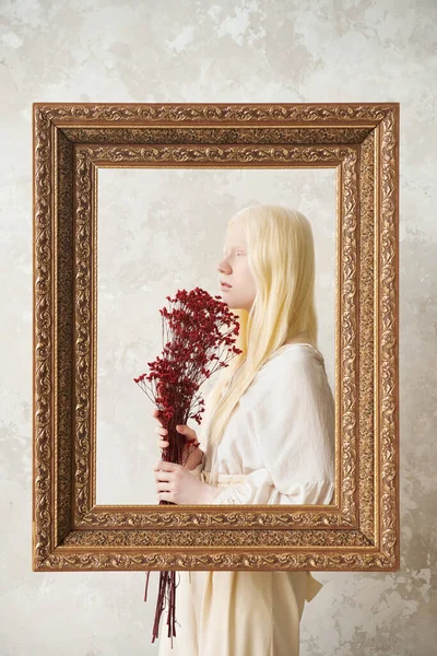 照片拍摄过程中 年轻漂亮的白化病妇女站在白色墙上的大画框后面 手里拿着一束干花 — 图库照片